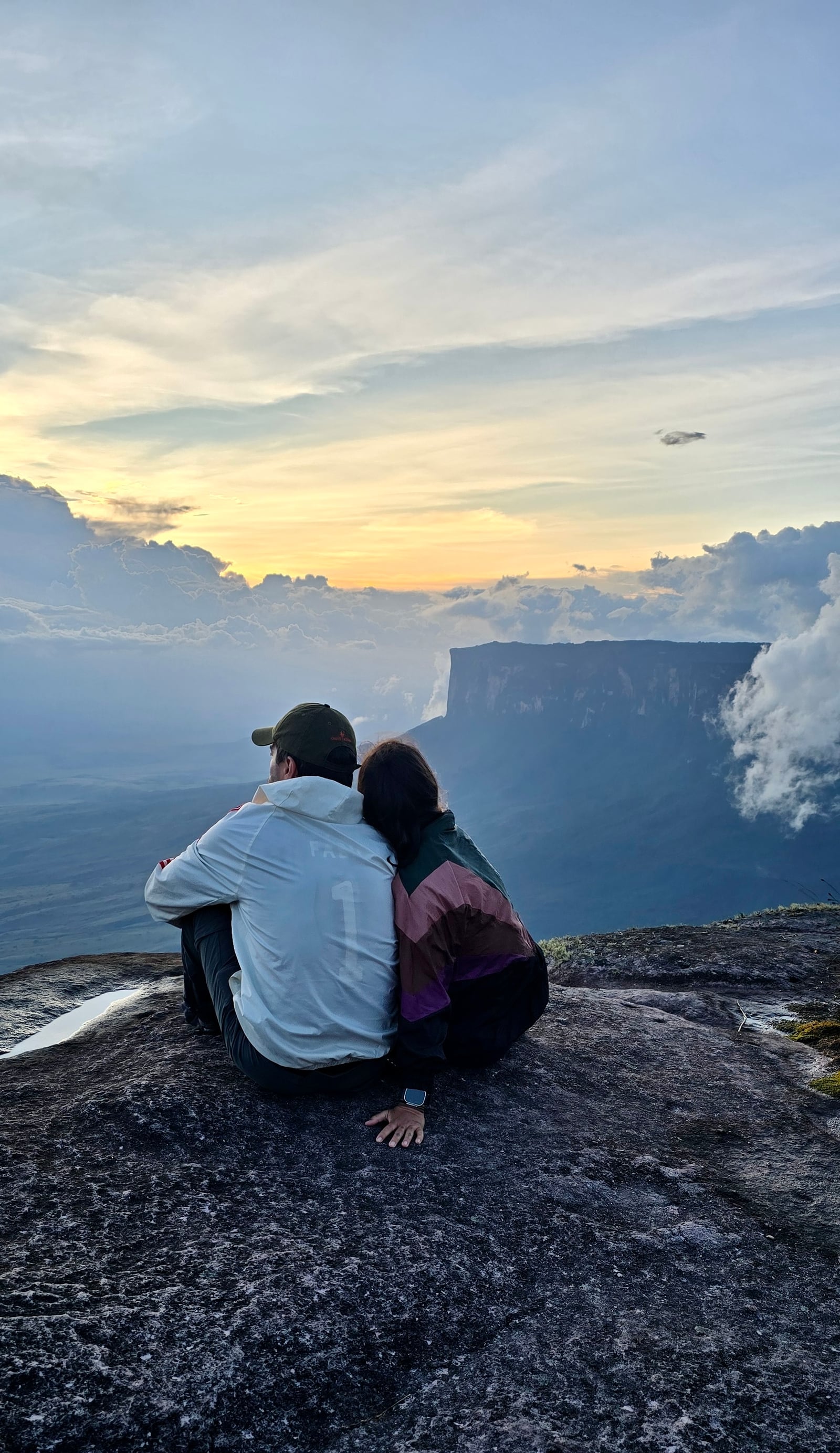 Thaíssa e Julián sentados de costas na borda, olhando a parede do tepui na neblina