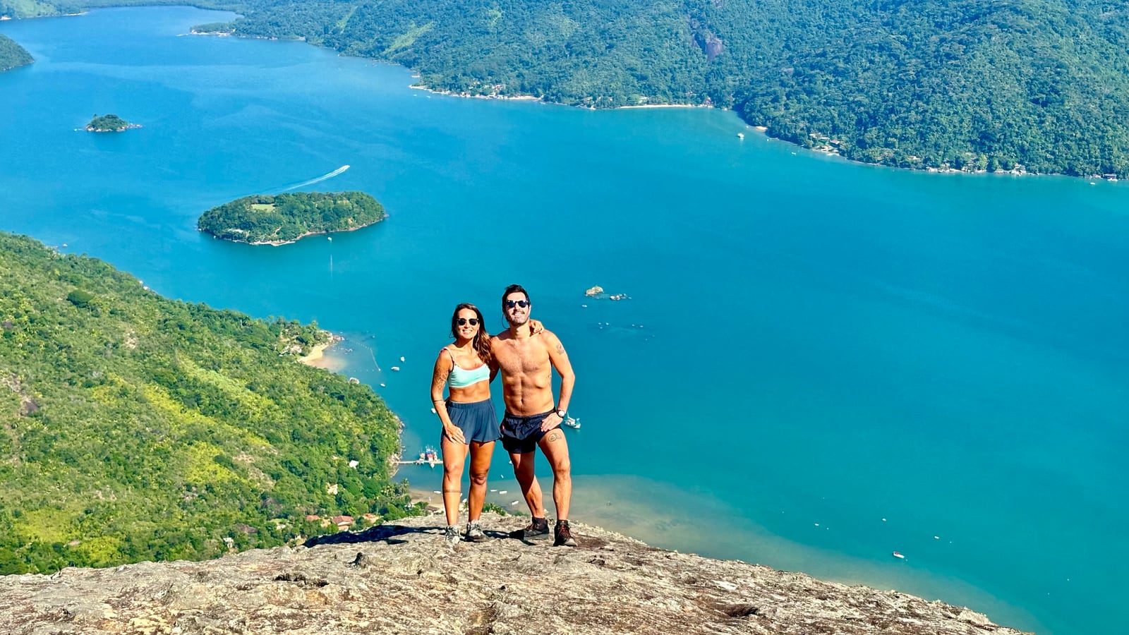 Thaíssa e Julián no alto de uma pedra, com a baía verde-azulada e as ilhas lá embaixo